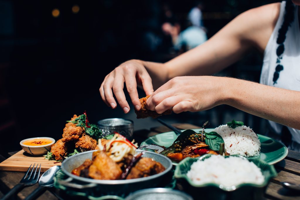 A woman is holding a piece of fried chicken on a plate filled with food.