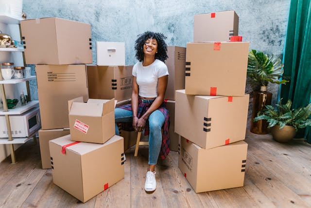 A smiling young woman sits in a chair surrounded by stacked moving boxes, feeling at ease and ready to feel at home in Nashville's new apartment.