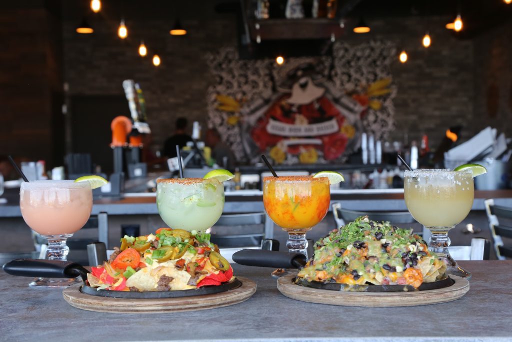 A table at Nacho Daddy Nashville featuring two sizzling plates of gourmet nachos alongside four colorful margaritas, with a lively bar and neon-lit décor in the background.