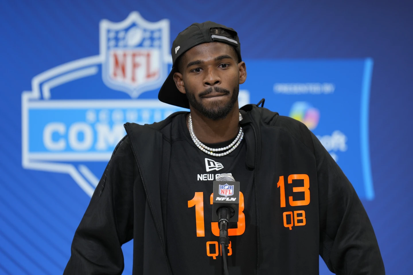 Shedeur Sanders, wearing a black jacket and hat, stands at a podium with an NFL microphone during the NFL Scouting Combine, with a focused expression on his face.