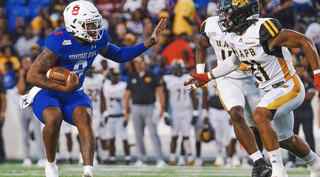 A Tennessee State University player in a blue uniform stiff-arms a University of Arkansas at Pine Bluff defender wearing a white and gold uniform during a football game at the 2023 Southern Heritage Classic. A crowd of spectators is visible in the background.