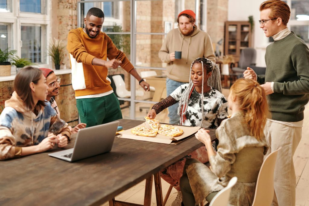 Diverse group of people eating pizza, looking at a laptop, laughing, and discussing