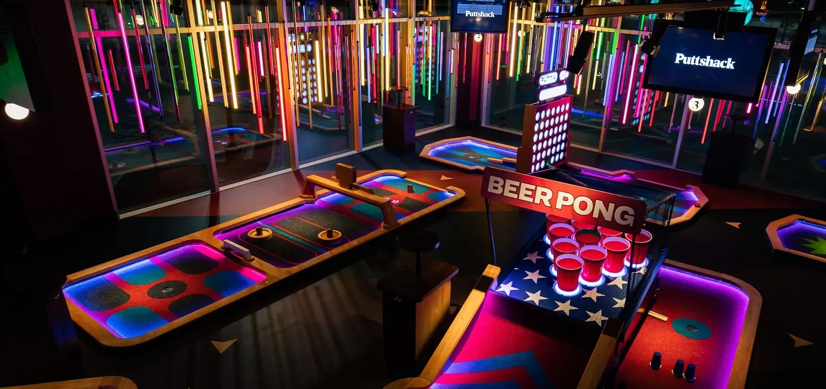 Interior of Puttshack Nashville featuring neon-lit mini-golf holes, colorful hanging lights, and a beer pong golf setup.