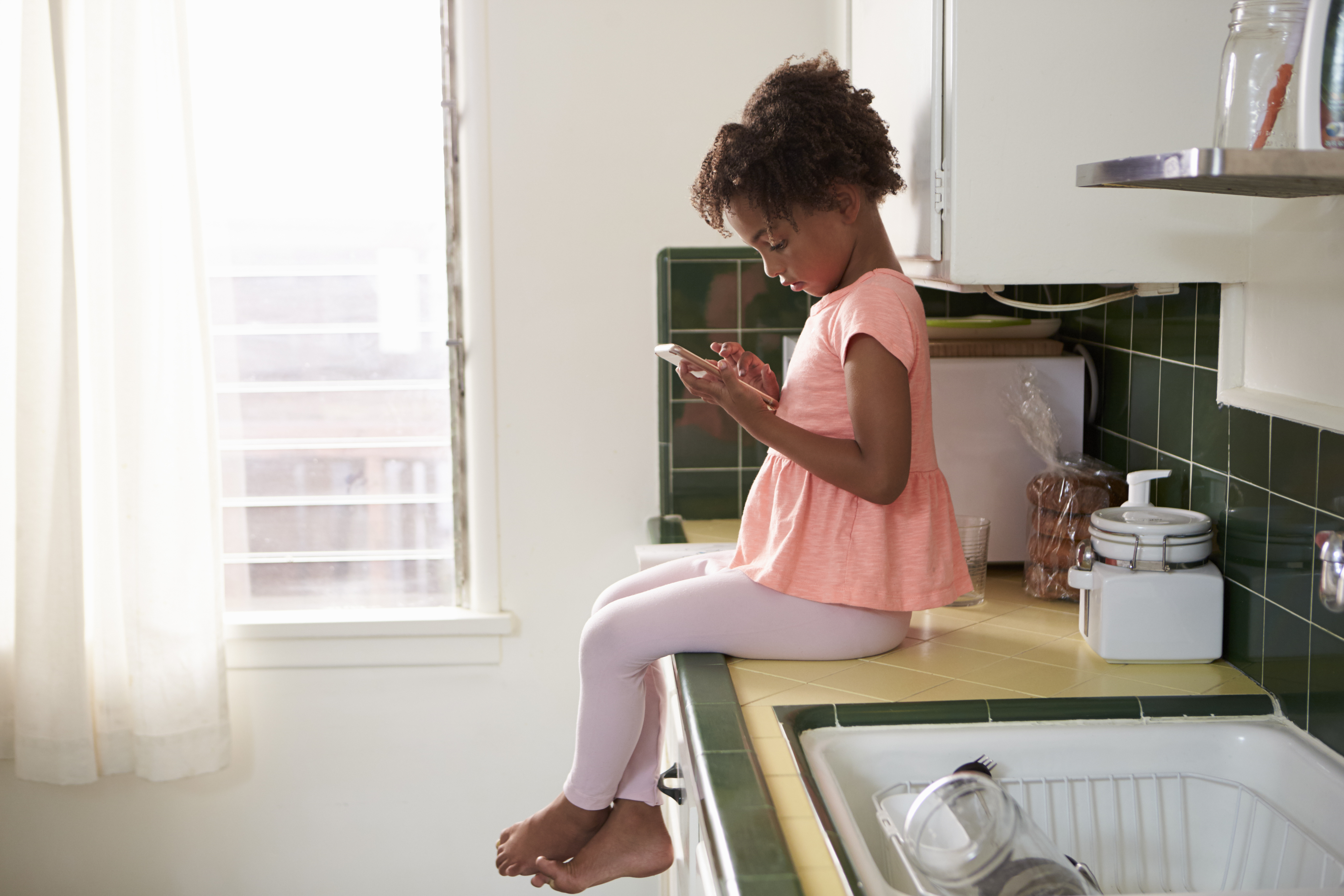 Young Girl Sits In Kitchen And Plays With Mobile Phone. (Photo by: monkeybusiness)
